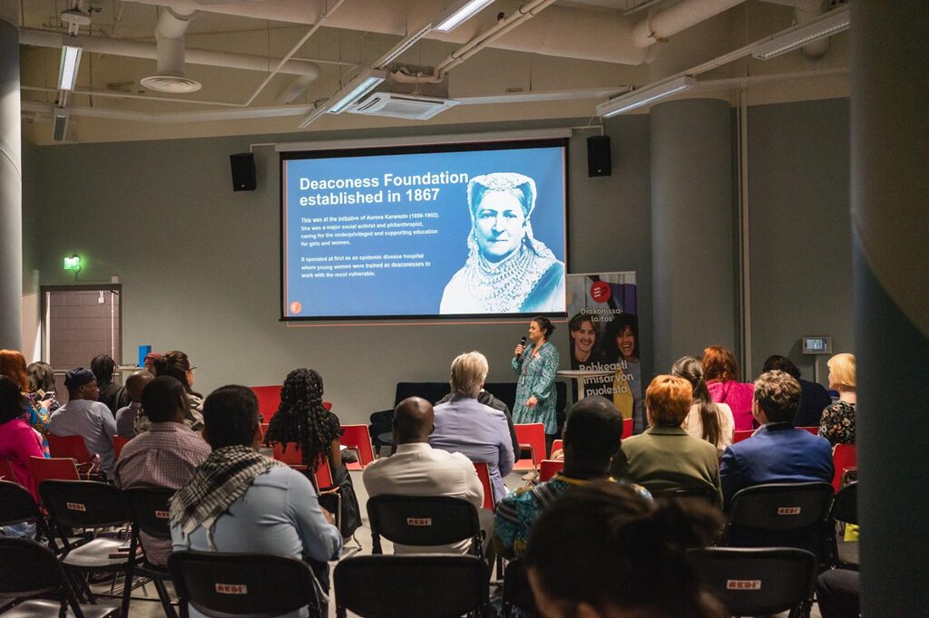 An audience is sitting and listening to a woman behind whom there is a large screen.