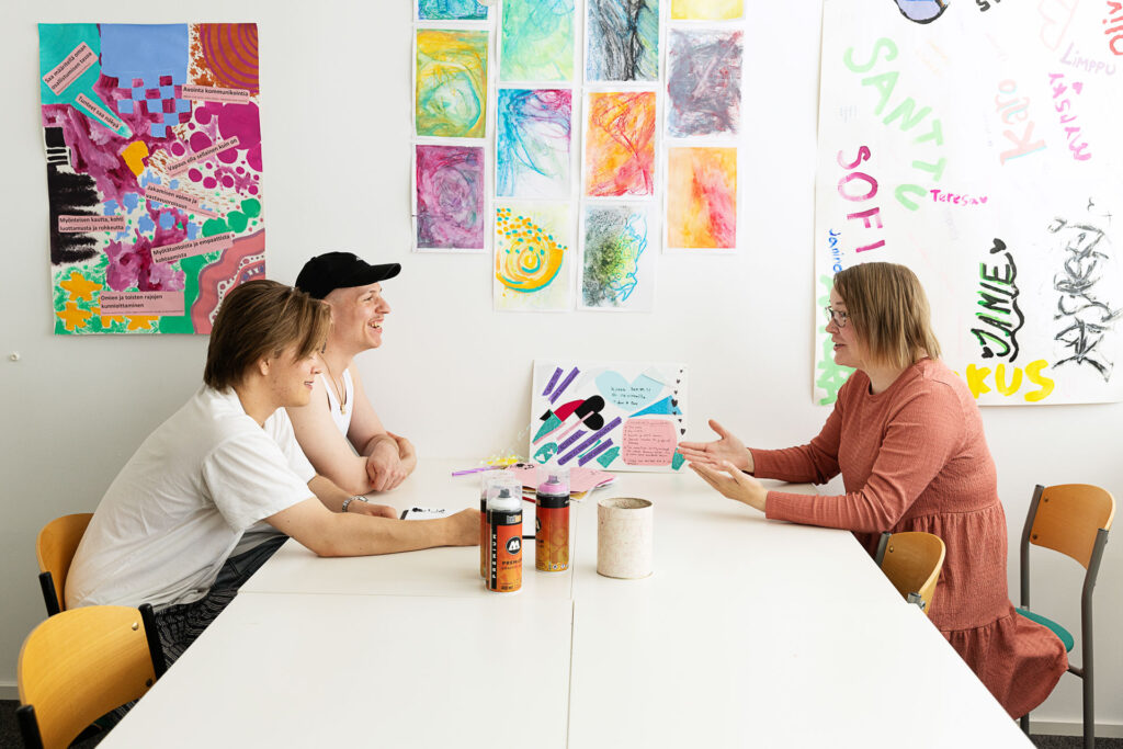 Kolme ihmistä istuu pöydän ääressä keskustelemassa. Taustalla seinälle on kiinnitetty värikkäitä taideteoksia.