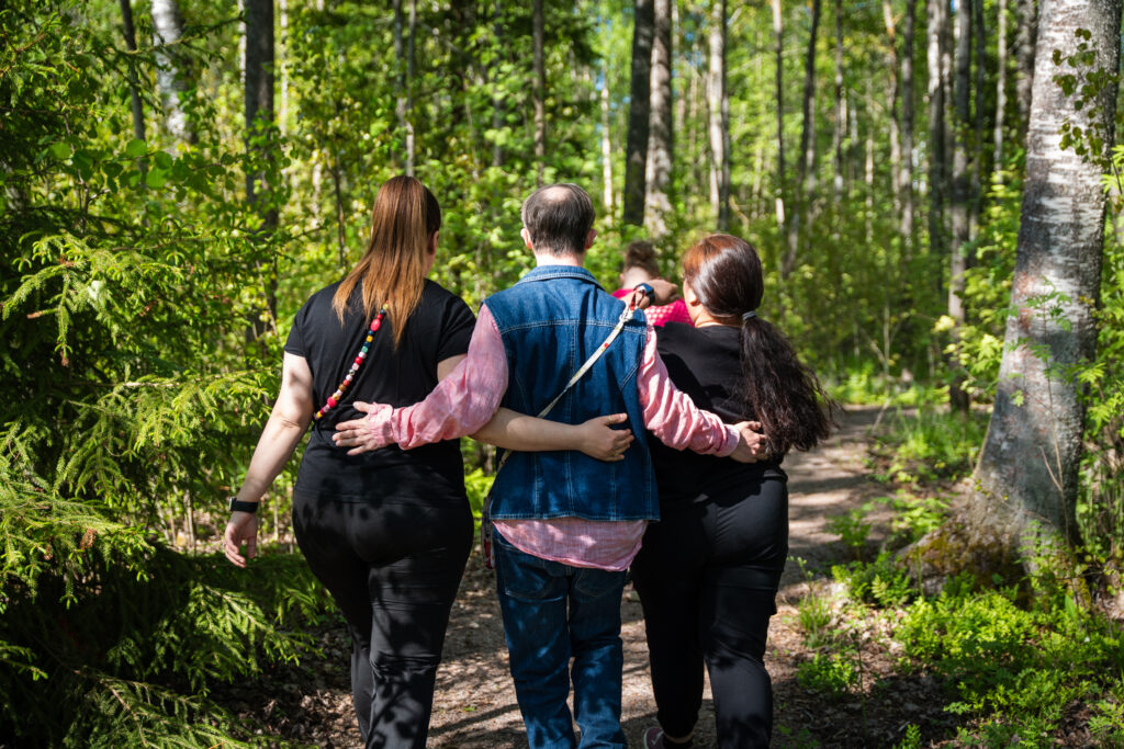 Kolme ihmistä kävelee metsässä vierekkäin toisistaan kiinni pitäen. Vammaistyö on myös tätä.