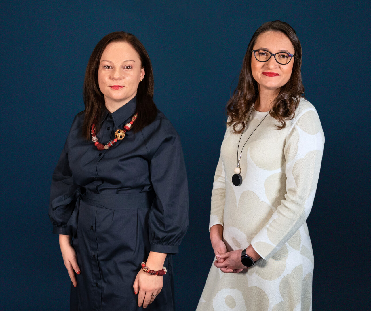 Two women are standing in front of a blue background looking at the camera.