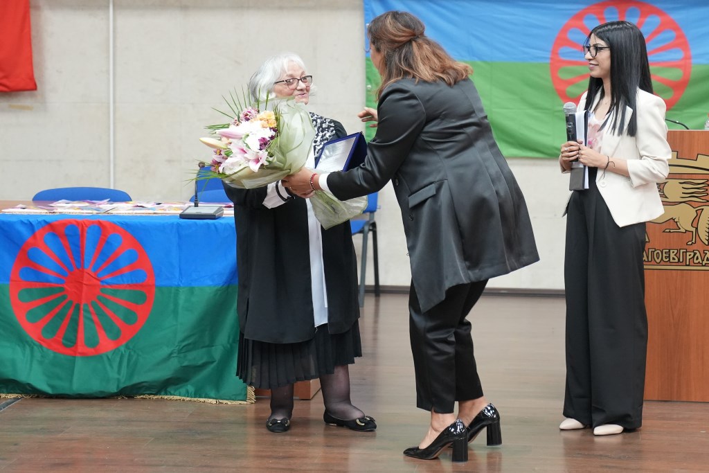 A white-haired woman receiving flowers from a younger one with Roma flags in the background.